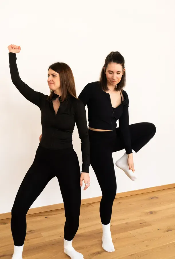 Zwei Trainerinnen in schwarzen Outfits bei einer verspielten Barre-Pose